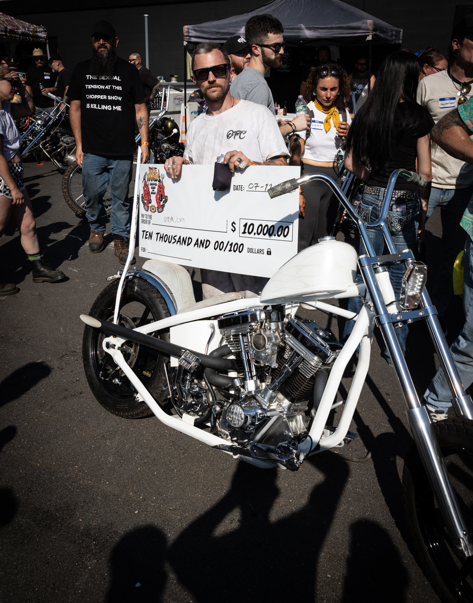 Idle Hands Chopper Show winner doom_cvlt holding a $10,000 check next to his white chopper Pale Horse